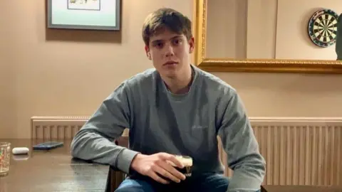David Wright David Wright sitting in a pub in front of a radiator and at a table while wearing a blue jumper and holding a Guinness. He is looking directly into the camera.