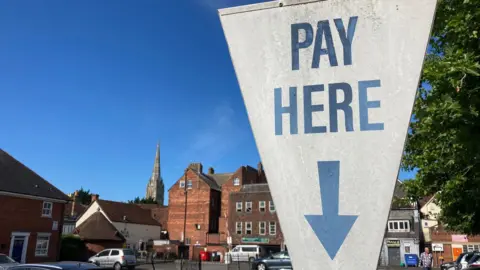 A white sign at a car park in Salisbury, Wiltshire, which reads "pay here" in blue writing, with a blue arrow pointing down. The sign is shaped like an upside down triangle. Cars can be seen parked in the city car park behind, which is surrounded by older buildings and a large spire can be seen in the background. It is a sunny day.