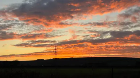 WeatherWatchers/Corvus A bright orange sunrise captured in Pewsey. The hillside in the foreground is completely in darkness with an electricity pylon highlighted by the cloudy orange sky beyond.