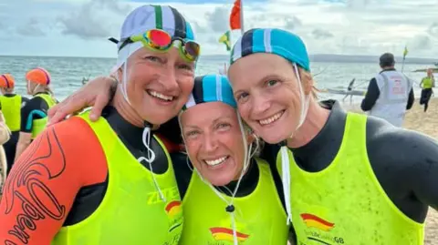 Gyllyngvase Surf Life Saving Club Three female surf lifeguards cuddled together smiling at the camera with the beach and sea behind them. They are wearing wetsuits, hi-viz vests and swim caps.