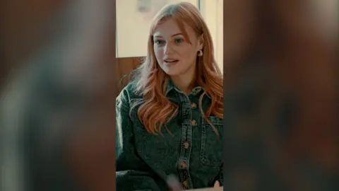 A woman with long wavy pink hair is talking to someone off camera. She is wearing a denim jacket with buttons.