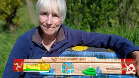 Lesley Sleight Lesley Sleight, wearing a blue jumper, leans on a table where the baton rests. It is a three-sided wooden baton, featuring coloured paintings of Manx heritage sites, and cultural events. 