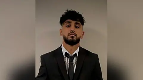 Uzair Khan with black hair and beard wearing a black suit and tie with a silver chain necklace.