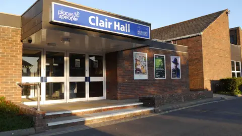 An exterior view of Clare Hall - a brown building with a blue sign above the door.