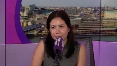 A woman speaks into a microphone against a backdrop of a city skyline and purple.