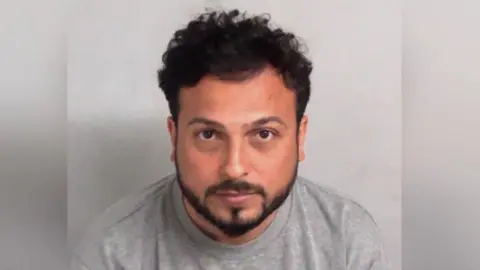 Essex Police A man looks directly into the camera. He has curly dark hair and a neat beard. He wears a grey top and is not smiling