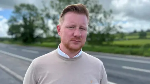 Daniel McCrossan stands on the side of a road with a neutral expression on his face as he looks at the camera. McCrossan has ginger hair which is styled and has a ginger beard. He is wearing a beige jumper with a white collared shirt underneath. Behind him is an empty main road and some trees, but they are blurred.
