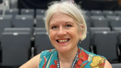 Harriet Heywood/BBC Ms Chantal is sat in the theatre seating smiling at the camera. She has medium length blonde hair, blue eyes and pink lipstick. She is wearing earrings and blye, red and gold flower print dress. She is also wearing glasses on top of her head. 