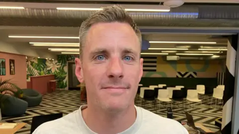 A man with short hair and blue eyes wearing a white t-shirt.