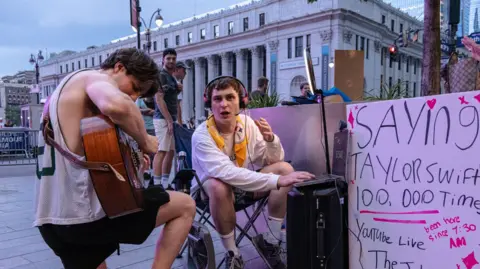 EPA A man playing a guitar kneels next to a man sat in a chair alongside a sign that reads: "Saying 'Taylor Swift' 100,000 times". 