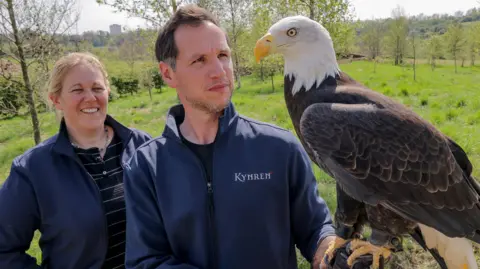 Kynren Jimmy, who is tall with faint grey stubble and short dark hair stands in a field holding a large brown eagle with a white head and yellow beak. Anna, who has blond hair, is standing smiling beside him.
