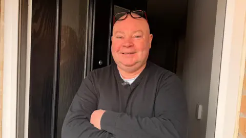 Jasmine Lowe/BBC News A bald man wearing a black jumper and red-framed glasses on the top of his head. He has bright blue eyes and is smiling. He has his arms folded and is standing in his front doorway looking at the camera. 