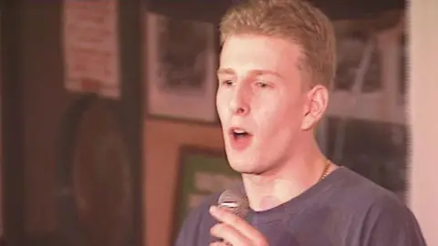 Paddy Kielty holding a silver microphone, wearing a navy top and gold necklace