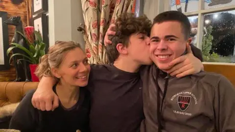 Kieron Duggan Tiger, wearing a black T-shirt, has his arms around his mother Lisa, who is blonde and wearing a black T-shirt, and his brother River who is wearing a grey hoodie. They are sitting in the booth of a restaurant. 