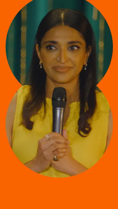 Sindhu Vee wears a yellow top and holds a microphone