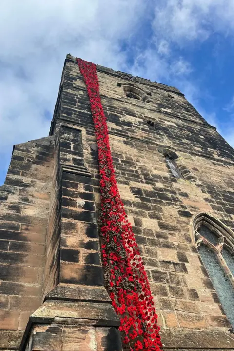 A beige and black coloured brick church that stretches into the sky. There is a long net attached to the top of the church and dangles to the bottom. Attached  to it are thousands of red and black knitted poppies