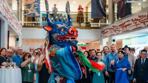 A person wearing a blue robe and a blue model head of a dragon, dancing in front of a crowd of people in a museum. 