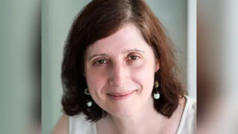 Cambridge University Hospitals Headshot of Dr Effrossyni Gkrania-Klotsas. She is smiling while wearing a white sleeveless blouse and matching white earrings. She has shoulder length brown hair and brown eyes.