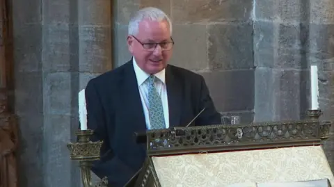 Dunblane Cathedral A man with short grey hair and glasses is wearing a black jacket, white shirt and green tie as he stands at a church lectern