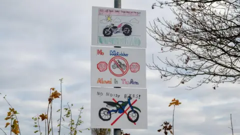 Three white signs on a metal pole. The top sign depicts a bike with big black tyres driving on grass. The sign below it has a man riding a bike with a red line through it and two red 'stop' signs. The bottom sign has a bike with a red cross drawn over it