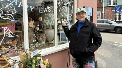 Juliet is wearing a cap and a dark coat.
She is standing next to a shop with a white, floral window mural that she painted on it.
The shop is on the High Street and there are flowers next to it.