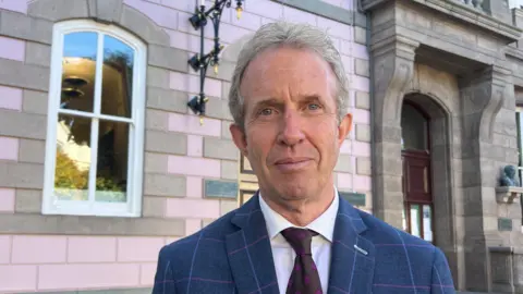 Deputy David Warr looks at the camera. He is wearing a maroon tie with pink spots, and a blue and pink tartan blazer. He is stood outside a stone building.