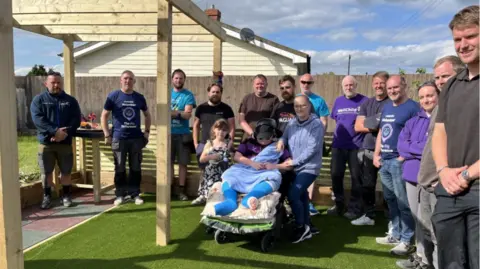 WellChild About a dozen people, mainly men in DIY work clothes, are pictured standing smiling to camera in a small garden. In the middle of them is a child in a wheelchair. The child has both legs in plaster and is wearing a baseball cap and sunglasses. There is a wooden pergola to the left.