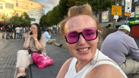 Martin Heath/BBC Close-up of Emma Gough  wearing large pink-tinted sunglasses and a white sleeveless top, seated in a town-centre public space and facing the camera. Other people are sitting and standing nearby, including one person on a phone and another seated with their back to the camera. Trees line the square, while shops and a modern building are visible in the background. The image is lit by warm evening sunlight.