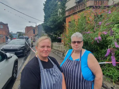 Cafe workers Joanne and Barbara standing at the cordon in front of the fire- damaged building. Joanne has blonde hair tied back and is wearing a navy t-shirt and a blue and white striped apron. Barbara has short, grey hair and dark glasses and is wearing a sleeveless blue top and a blue and white stripped apron. There is a shrub with large purple flowers growing out of the garden in front of the house.