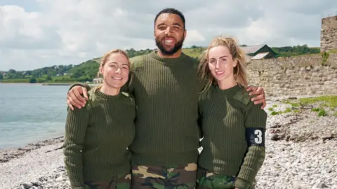 O canal 4 Lucy Spraggan, Troy Deeney e Michaella McCollum ficam em uma praia com os braços, sorrindo para a câmera. Os três estão usando saltadores de estilo verde do Exército e calças de camuflagem.