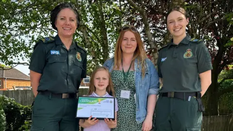 Kathleen Rule, Jessica, Rhian and Kiya Wood stand in a residential garden. Kathleen and Kiya are wearing paramedic uniform. Jessica is wearing school uniform and her mother Rhian is in a denim shirt and green dress underneath.
