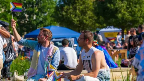 Two men holding Pride flags at a Pride in Gloucestershire Event in 2025. It is a sunny day and they are sitting down. There are lots of people milling about around them.