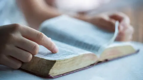 An open bible on a table. A person's hands are visible, turning the pages.