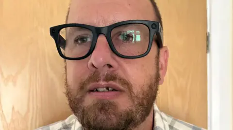 Contributed A close up of a man's face who is wearing smart glasses. He has short hair and a beard and there is a wooden door behind him.