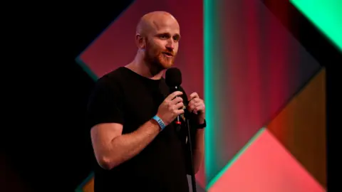 Ray Bradshaw - a bald man with a ginger beard, holding a microphone while performing stand up comedy onstage.