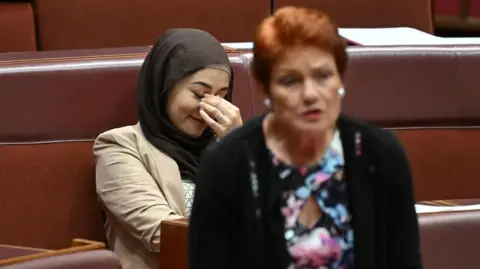 Reuters A woman in a headscarf touches her fingers to her face behind a woman with short red hair who is speaking. 