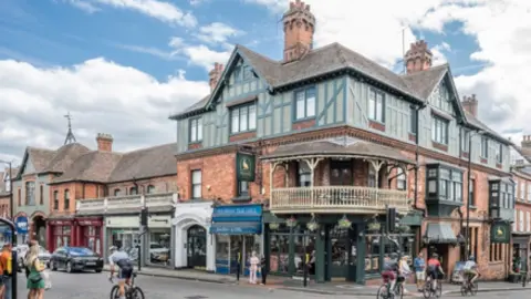Ricky Barnett The Peahen is a large corner building on a junction. It has a small curved balcony on its corner over the entrance to the pub. The image also shows neighbouring shops, cyclists on the road and people waiting to cross the road at a crossing.