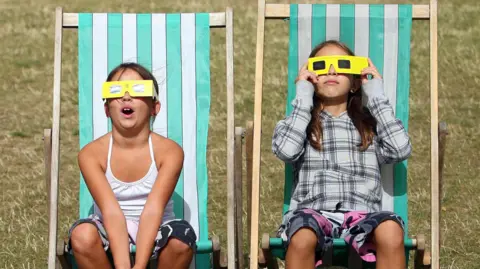 Two girls are sitting on green and white stripy deck chairs on grass wearing yellow eclipse glasses. They're looking up and it is a bright day.