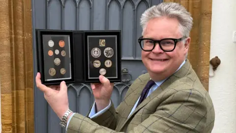 A smiling Richard Bromell holding up a black case containing a set of 14 commemorative coins. He has short grey hair and is wearing black-rimmed glasses and a moss green windowpane-tweed jacket.