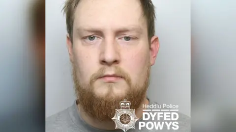 Dyfed-Powys Police Police mugshot of Rhys Jamieson. He has a droopy left eye, scruffy hair and beard, grey t-shirt