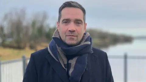 Michael Cadden looks into the camera wearing a large black and brown scarf and dark overcoat. He has black hair with grey coming through. He is standing in front of a body of water with guard railings around him.