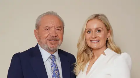 A woman with long blonde wavy hair wearing a white jumpsuit stands next to a man with a tanned face, short grey hair and stubble. He's wearing a suit. They're both looking at the camera and smiling.