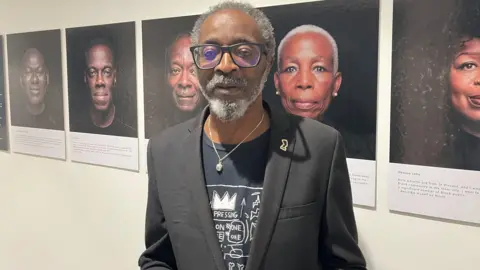 Don John stands in front of portraits displayed in the exhibition. He is wearing dark-framed glasses and has white hair and a white and grey beard. He is wearing a charcoal grey blazer with a small gold brooch on his top left lapel over a black t-shirt with a white design visible on the front. He is also wearing a silver chain around his neck with a small pendant that hangs in front of his t-shirt.