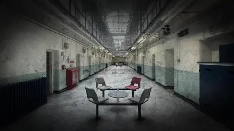 BBC Four chairs and a small table are placed in the long hallway of Shepton Mallet prison. There is a net which separates the ground floor with the other floors.