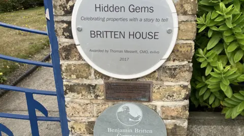 GUY CAMPBELL/BBC Grey round signs outside the property on a brick will reading "Hidden Gems - Britten House" 