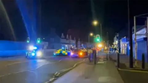 Phone image of the crash scene with two police cars with their lights flashing parked by a pelican crossing with an abandoned car on it