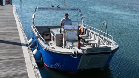 The 12 seat ferry tied to a pontoon on flat calm water. It's open topped and blue coloured