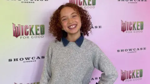 Esme smiling at the camera as she attends the screening, wearing a grey jumper with a blue collar. She has brown curly hair.