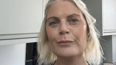 Heather Daly A selfie of a woman with bobbed white-blonde hair stands in a kitchen with pale grey cabinets behind her. She has blue eyes and is half-smiling with her mouth closed.
