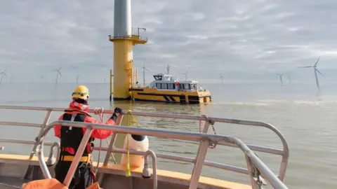 Monty Rakusen/Getty Images An engineer standing on the deck of a boat wearing orange and black clothing as another boat approaches one of a number of offshore wind turbines.